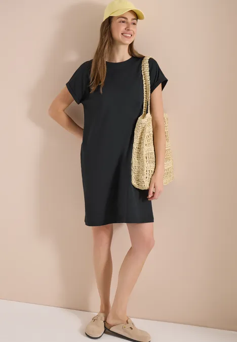 Vrouw in zwart T-shirtjurk met strooien tas en beige instapschoenen.