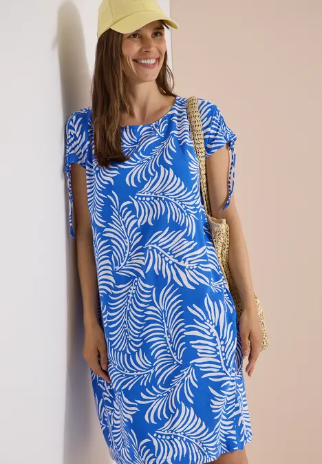 Knielanges Kleid mit Capsleeves surf blue Frau in blau-weiß gemustertem Kleid, Strohhut und Tasche steht an einer Wand.