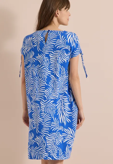Knielanges Kleid mit Capsleeves surf blue Frau von hinten in knielangem, blauem Kleid mit weißem Blattmuster, kurzen Ärmeln und seitlichen Schleifen.