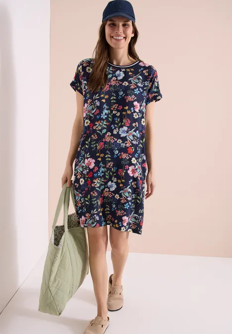 Frau trägt kurzärmeliges Kleid mit buntem Blumenmuster, Basecap, Stofftasche und Sneaker