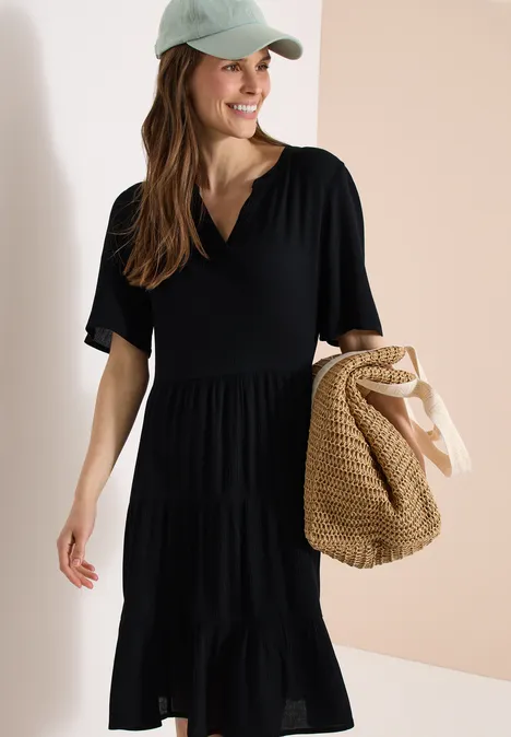 Vrouw in zwarte laagjesjurk met V-hals, draagt een lichte pet en houdt een gevlochten strooien tas vast.