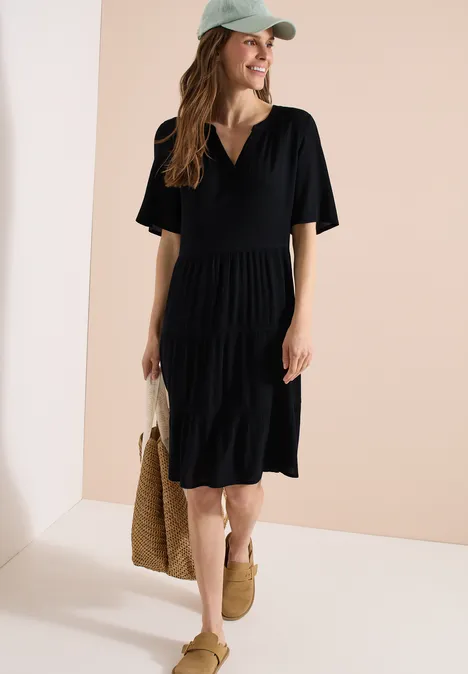 Vrouw in zwarte midi-jurk met V-hals en elastische taille, rieten tas, sandalen en pet.