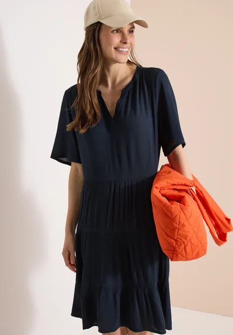 Frau in navyblauem Midikleid mit V-Ausschnitt hält eine orangefarbene Steppjacke, Gesicht nicht erkennbar.