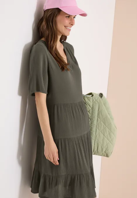 Vrouw in kaki wijd uitlopend jurkje met korte mouwen, met een gewatteerde schoudertas.