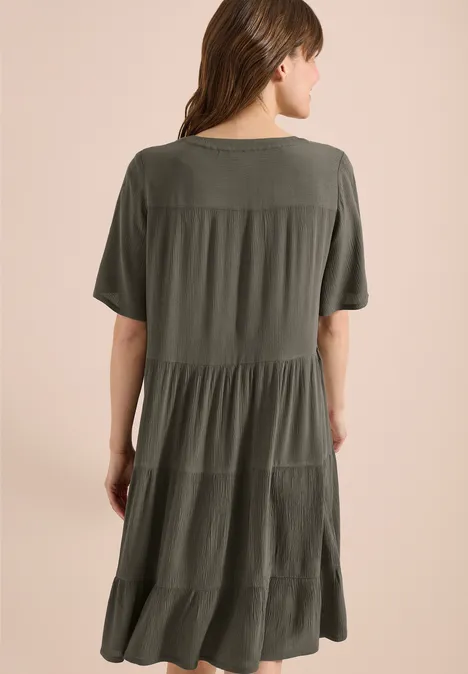 Vrouw in een losse, gelaagde midi jurk in olijfgroen met korte mouwen, rugaanzicht.