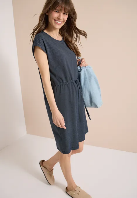 Vrouw in een knielange donkerblauwe jurk met korte mouwen en espadrilles, houdt een lichtblauwe trui vast.