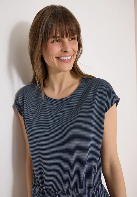 Een slanke vrouw voor een witte muur draagt een donkerblauw T-shirt en oorbellen.