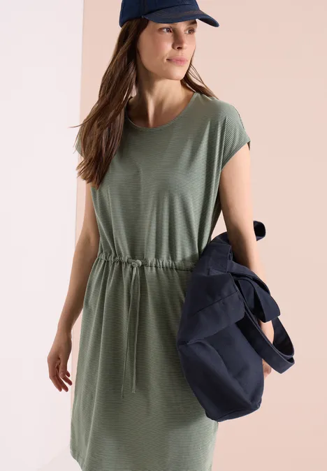 Vrouw in olijfgroene, kort mouwige jurk met taillekoord, die een donkerblauwe tas vasthoudt.