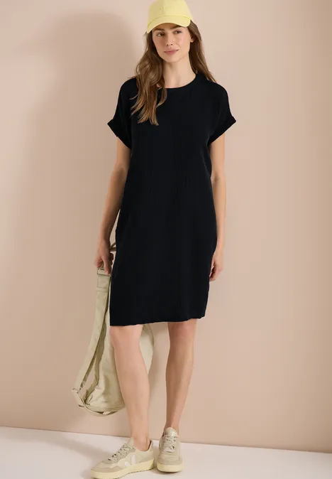 Frau in schwarzer T-Shirt-Kleid, Sneakers und Tasche vor neutralem Hintergrund.