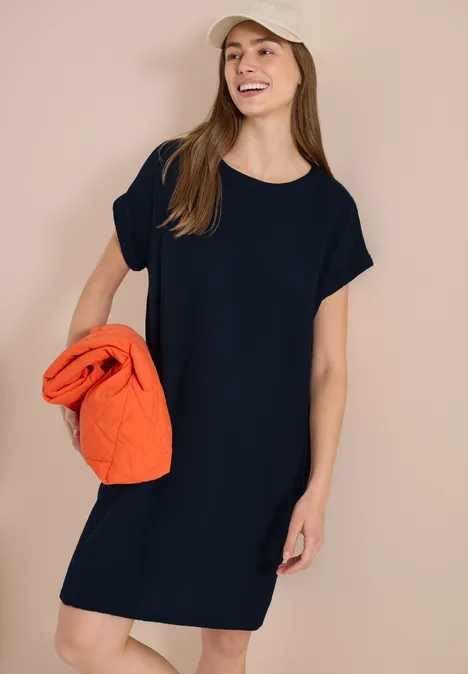 Frau trägt schwarzes, kurzärmeliges Kleid und hält orangefarbenen Pullover