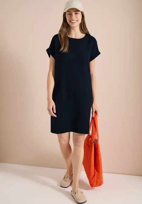 Frau in schlichtem schwarzen Kurzarmkleid, beigen Schuhen und einer orangefarbenen Tasche.