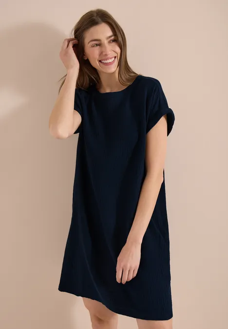 Frau in lockerem dunkelblauem T-Shirt-Kleid vor schlichtem beigem Hintergrund.