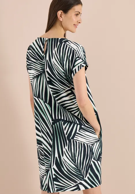 Vrouw voor een beige achtergrond in losse, korte mouwen jurk met groen-witte bladprint en zijzakken.