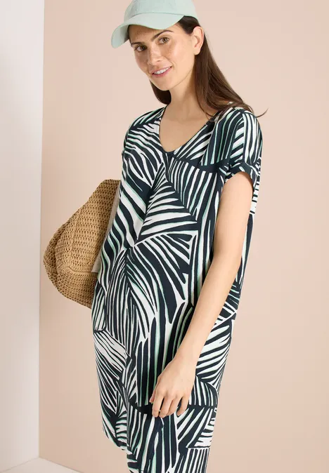 Vrouw in zomerse groen-witte bladprintjurk met strooien tas voor een beige achtergrond.