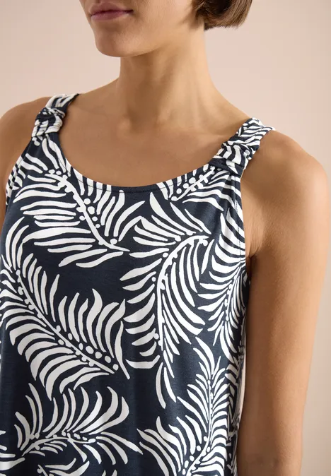 Close-up van een vrouw in een mouwloze, donkerblauwe top met wit bladmotief, schouders en hals zichtbaar.