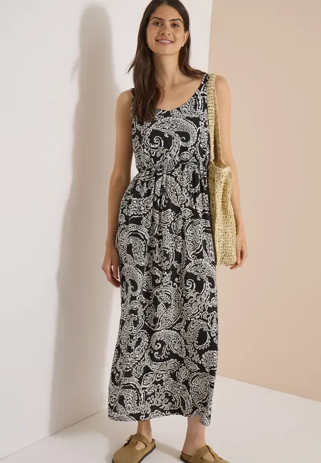 Frau in ärmellosem, schwarz-weißem Maxikleid mit Paisley-Muster trägt Korbtasche und Sandalen vor hellbeigem Hintergrund