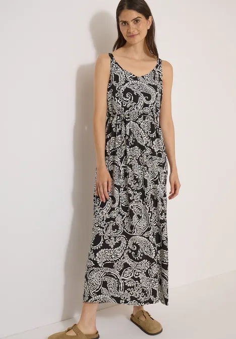 Eine Frau trägt ein ärmelloses Maxikleid in Schwarz-Weiß mit Paisley-Muster und olivgrünen Slippern.