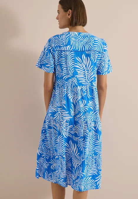 Eine Frau steht mit dem Rücken zur Kamera und trägt ein blau-weißes, kurzärmeliges, knielanges Kleid mit Blattmuster.