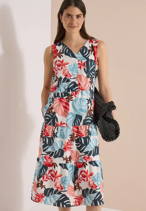 Femme en robe d'été sans manches à volants, à imprimé fleuri et feuillage coloré, tenant un sac noir en crochet.