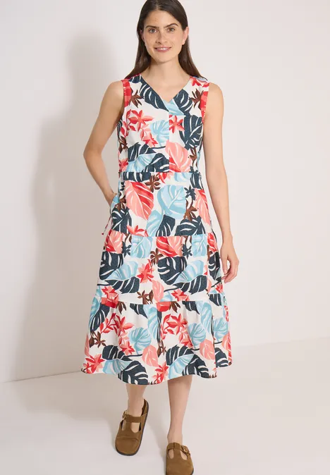 Femme en robe midi sans manches blanche à motif floral rouge et bleu, chaussures brunes, devant un fond clair.