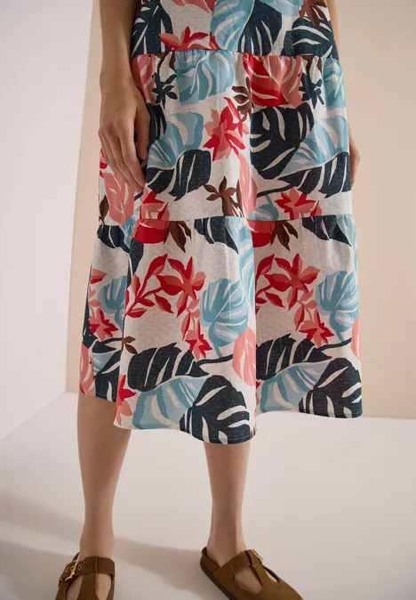 Jambes en sandales marron et robe blanche mi-longue ornée de larges feuilles rouges, bleues et vertes.