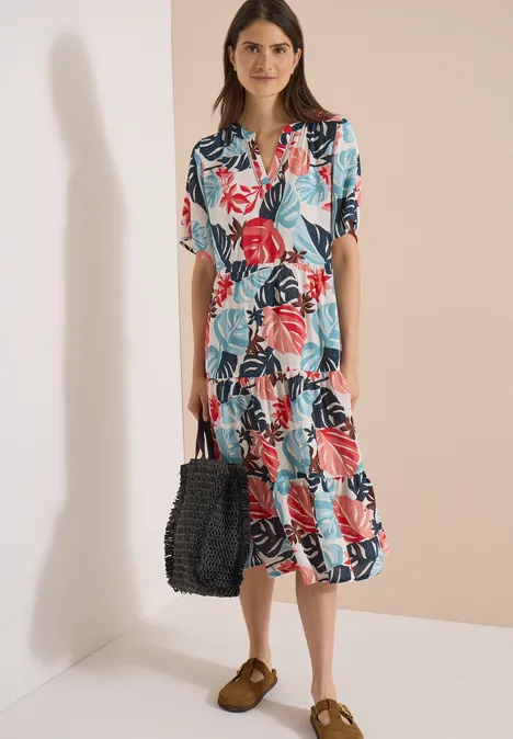Vrouw in midijlange jurk met kleurrijke bloemen- en bladprint, korte mouwen, met gevlochten tas en loafers.
