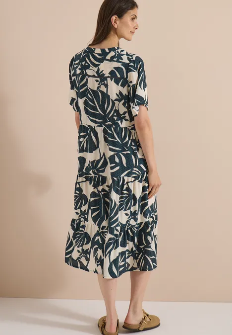 Robe mi-longue à col fendu et imprimé palm breeze green Femme de dos en robe mi-longue beige à imprimé feuilles bleues, sandales.