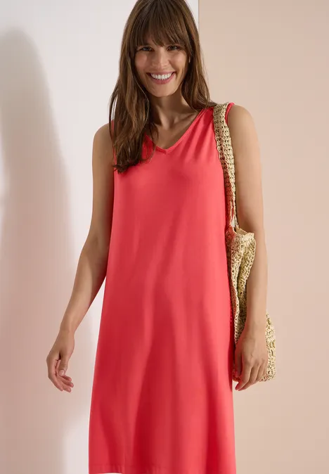 Knielanges Jerseykleid mit V-Neck coral Frau im ärmellosen Korallen-Sommerkleid mit geflochtener Tasche vor hellem Hintergrund.