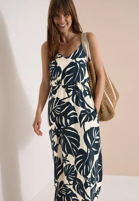 Maxikleid mit V-Neck und Print palm breeze green Frau in ärmellosem Maxi-Kleid mit großem Blattmuster und geflochtener Strohtasche vor neutraler Wand.