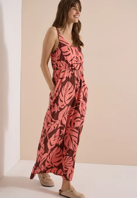 Maxikleid mit V-Neck und Print coral Frau im ärmellosen roten Jumpsuit mit schwarzem Blattmuster und Sneakers vor hellem Hintergrund.