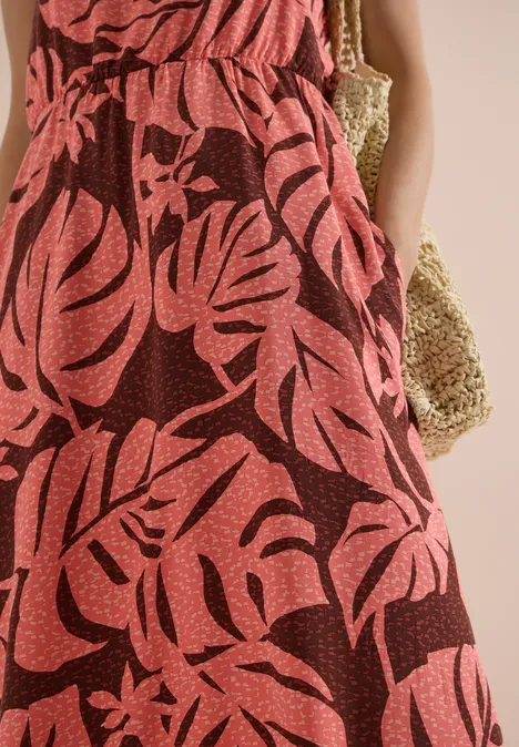 Maxikleid mit V-Neck und Print coral Rotes Maxikleid mit tropischem Blattprint, Frau mit Händen in den Taschen und Basttasche am Arm.