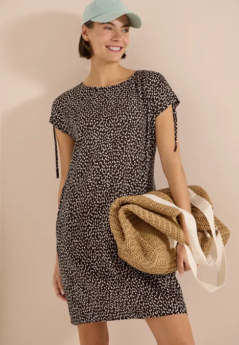 Frau in gemustertem Kurzarmkleid, mit strohfarbener Strandtasche und Baseballcap.