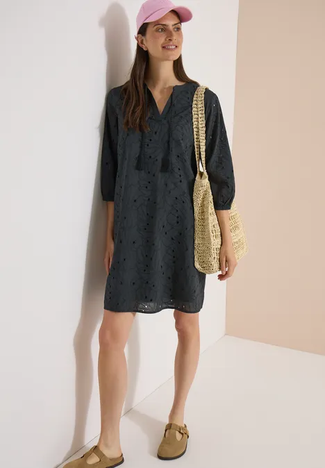 Vrouw in marineblauwe, lange mouwen jurk met opengewerkt patroon, strooien tas en pet.