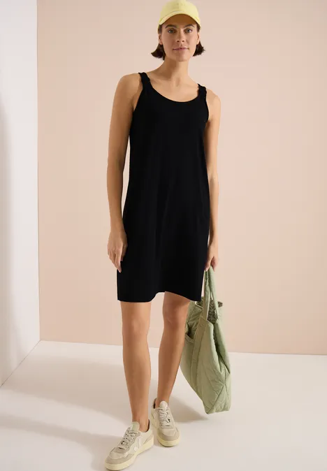 Zwarte mouwloze jurk, lichte sneakers, groene gestreepte tas, minimalistische achtergrond.