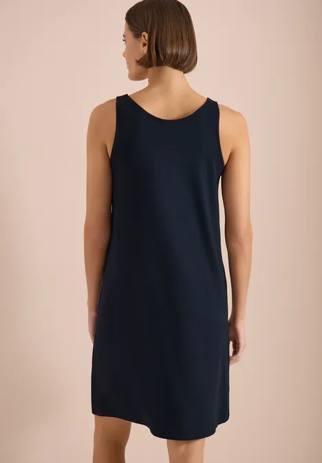 Vrouw van achteren in een mouwloze, knielange marineblauwe jurk tegen een beige achtergrond.