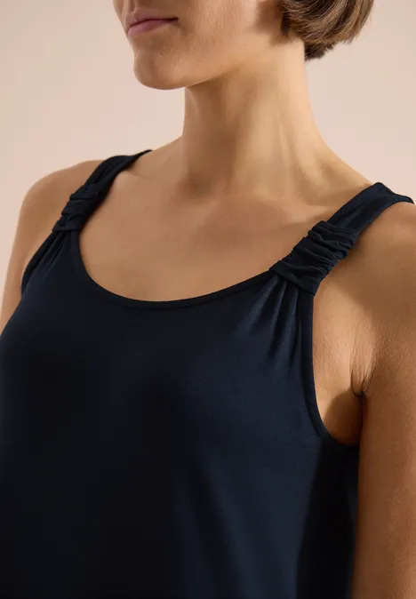 Vrouw in marineblauw mouwloos topje met geplooide bandjes, gedeeltelijke weergave tegen lichte achtergrond.