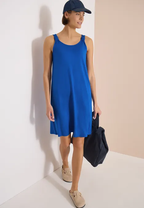 Knielanges Kleid mit Knotendetail surf blue Frau in lockerem, ärmellosem blauem Kleid mit Cap, hält eine dunkelblaue Tasche, trägt beiges Espadrilles-Paar.