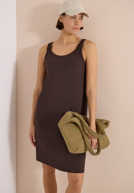 Vrouw in mouwloze bruine jurk, met pet en een groene tas in de hand.