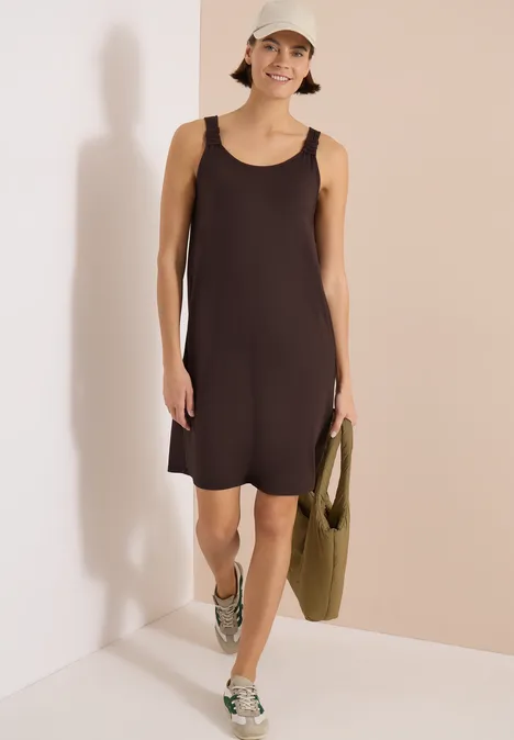Vrouw in donker mouwloze jurk en sneakers voor beiger wand, met groene tas.
