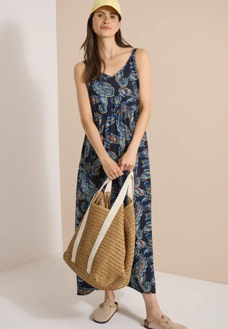 Maxikleid mit V-Neck und Print universal blue Frau im floralem Maxikleid mit Strohtasche und weißen Sneakers vor neutralem Hintergrund.