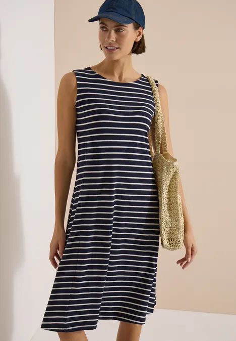 Femme en robe midi rayée bleu et blanc, sac en paille et casquette devant un mur clair.