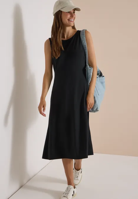 Frau im schwarzen ärmellosen Midikleid, weiße Kappe, blaue Tasche und Sneakers vor hellem Hintergrund.