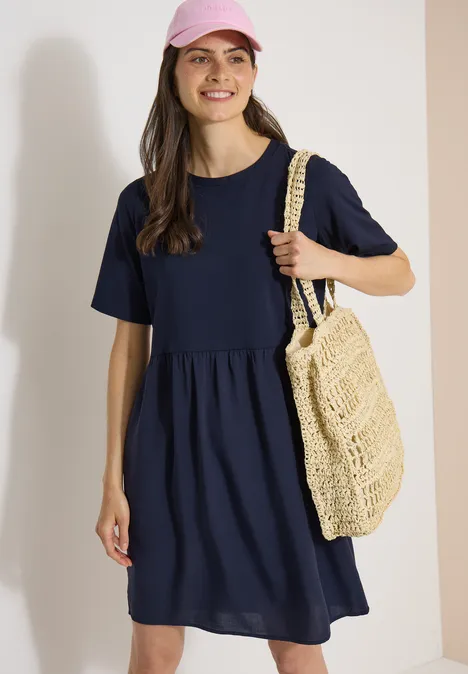 Knielanges Kleid mit Strukturmix urban dark blue Frau in lockerem dunkelblauem Kleid mit halblangen Ärmeln und gehäkelter Stroh-Tasche über der Schulter.