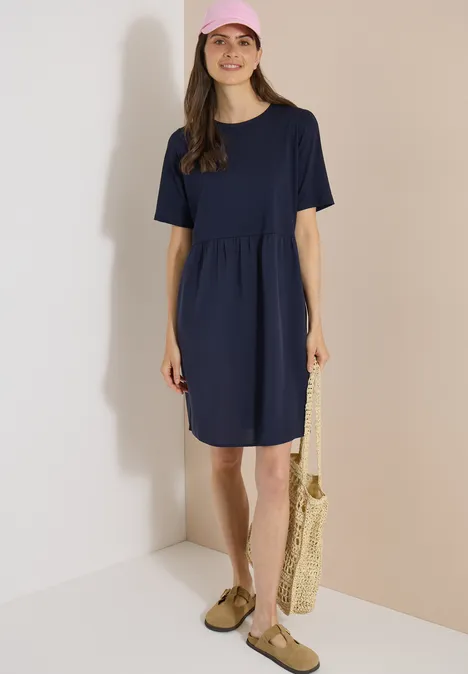 Knielanges Kleid mit Strukturmix urban dark blue Frau in knielangem dunkelblauem Kleid mit kurzen Ärmeln hält eine Basttasche vor neutralem Hintergrund.