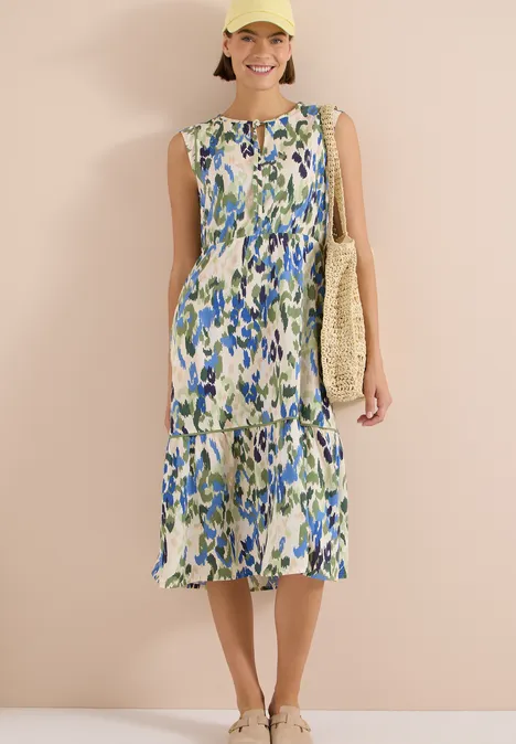 Midikleid mit Split Neck und Print cashmere beige Frau im ärmellosen Midikleid mit blau-grünem Blumenmuster und Strohtasche vor beiger Wand.