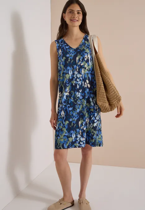 Frau im ärmellosen, knielangen Blumen-Sommerkleid in Blau-Grün mit geflochtener Tasche vor schlichtem Hintergrund.