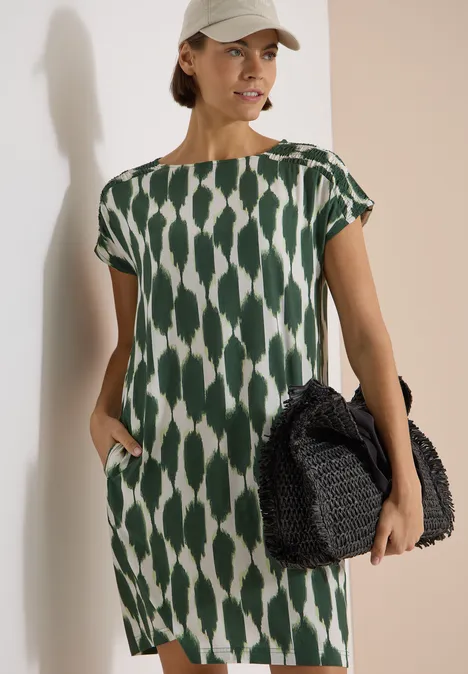 Knielanges Kleid mit Capsleeves und Print vanilla white Dame trägt ein locker sitzendes grün-weiß gemustertes Kleid mit kurzen Ärmeln und hält eine schwarze Flecht-Handtasche.