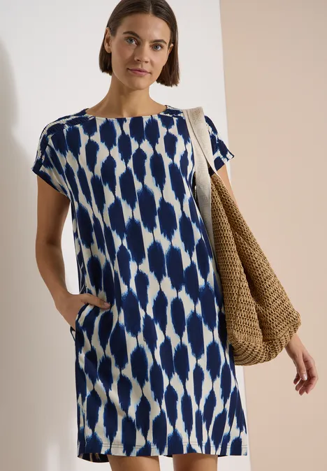 Knielanges Kleid mit Capsleeves und Print urban dark blue Frau im blau-weiß gemusterten knielangen Kleid mit Taschen und geflochtener Strohtasche.