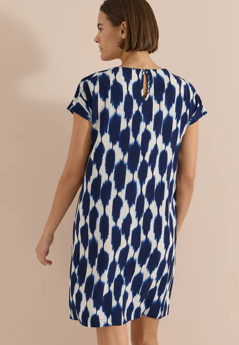 Knielanges Kleid mit Capsleeves und Print urban dark blue Frau von hinten in kurzärmeligem, weiß-blau gemustertem Shiftkleid vor beigem Hintergrund.