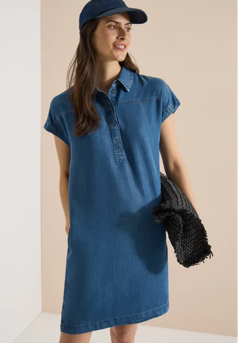 Frau in blauem Jeanskleid und Cap hält schwarze Tasche vor hellem Hintergrund.